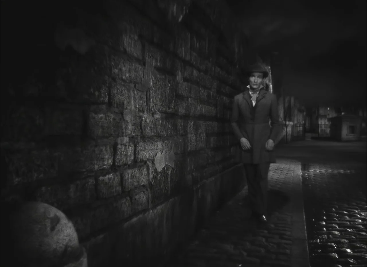 Les Enfants Du Paradis (1945) movie still: fullbody — A wet cobblestone street runs alongside a tall, weathered stone block wall at night. A lone…
