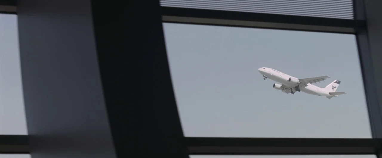 Syriana (2005) movie still: wide — A view through a modern airport terminal window shows a white commercial airplane in flight. The dark, thick…