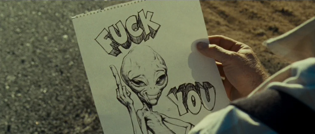 Paul (2011) movie still: closeup — A person holds a white piece of paper in an open desert environment. The paper features an ink drawing of an…