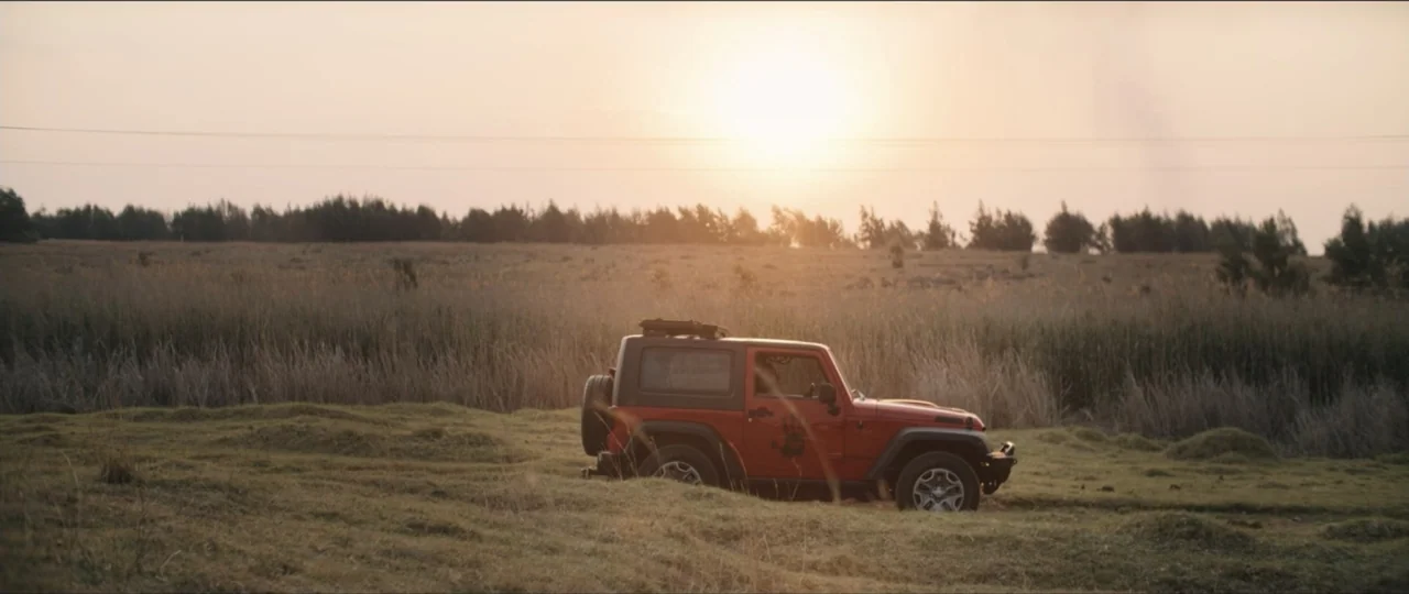 My Girlfriend's Father (2023) movie still: wide — A flat grassy field extends to a distant tree line under a bright sunset. A red two-door jeep is…