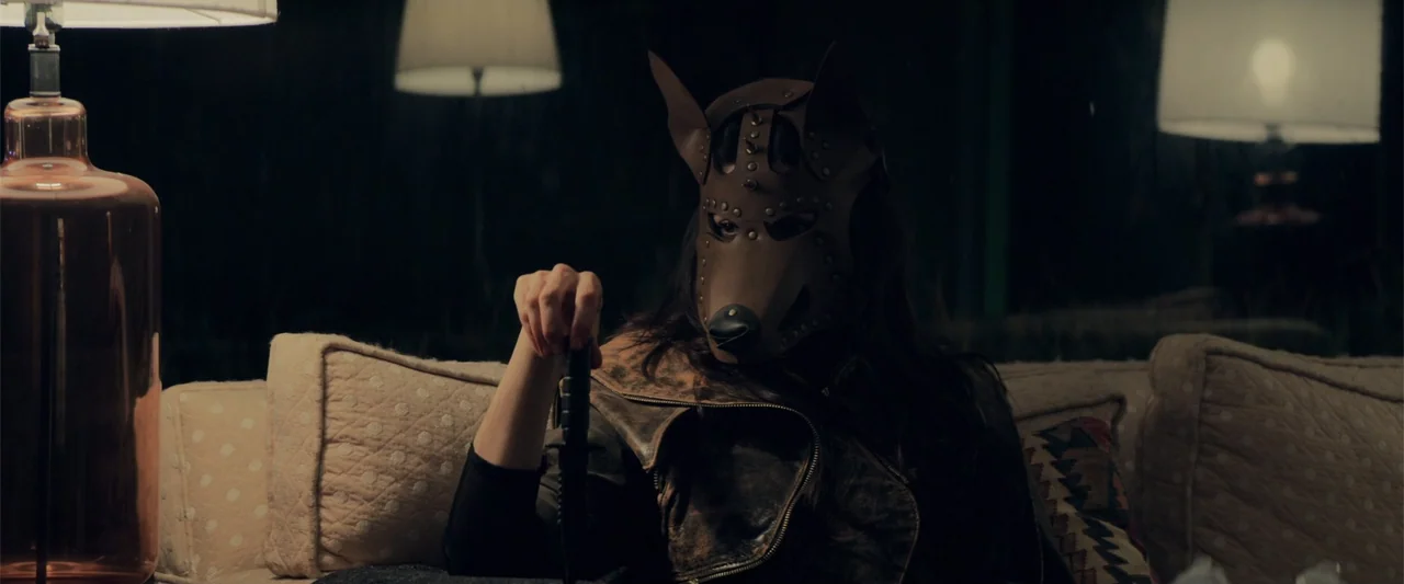 Human Animals (2020) movie still: medium — A dimly lit room features a person wearing a leather dog mask seated centrally. The subject grips the…
