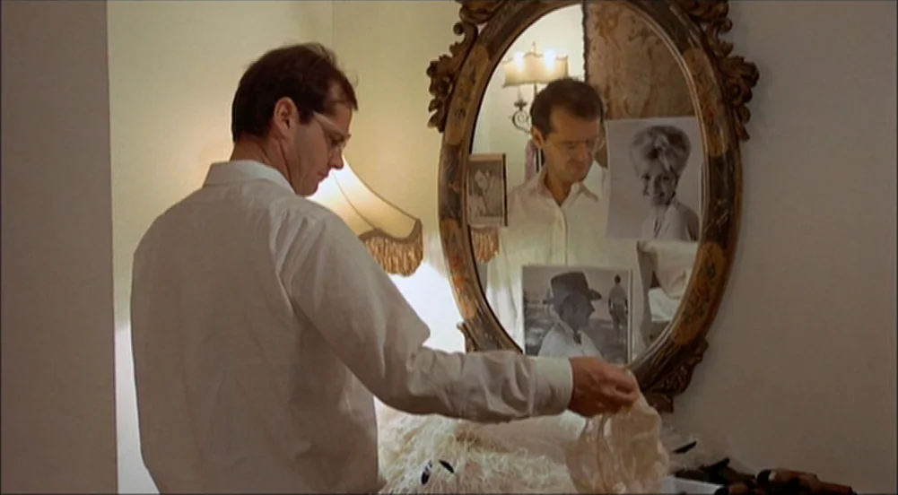 The King Of Marvin Gardens movie still: medium — A man stands in front of an ornate oval mirror mounted on a white wall. He examines a piece of…