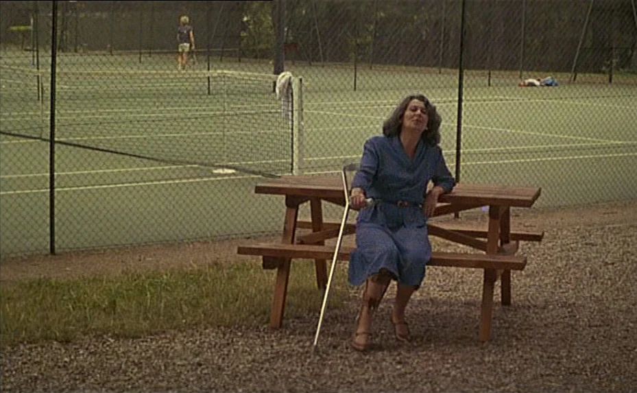The Woman Next Door (1981) movie still: fullbody — A woman sits on a wooden picnic table beside a fenced tennis court. She wears a blue dress and…