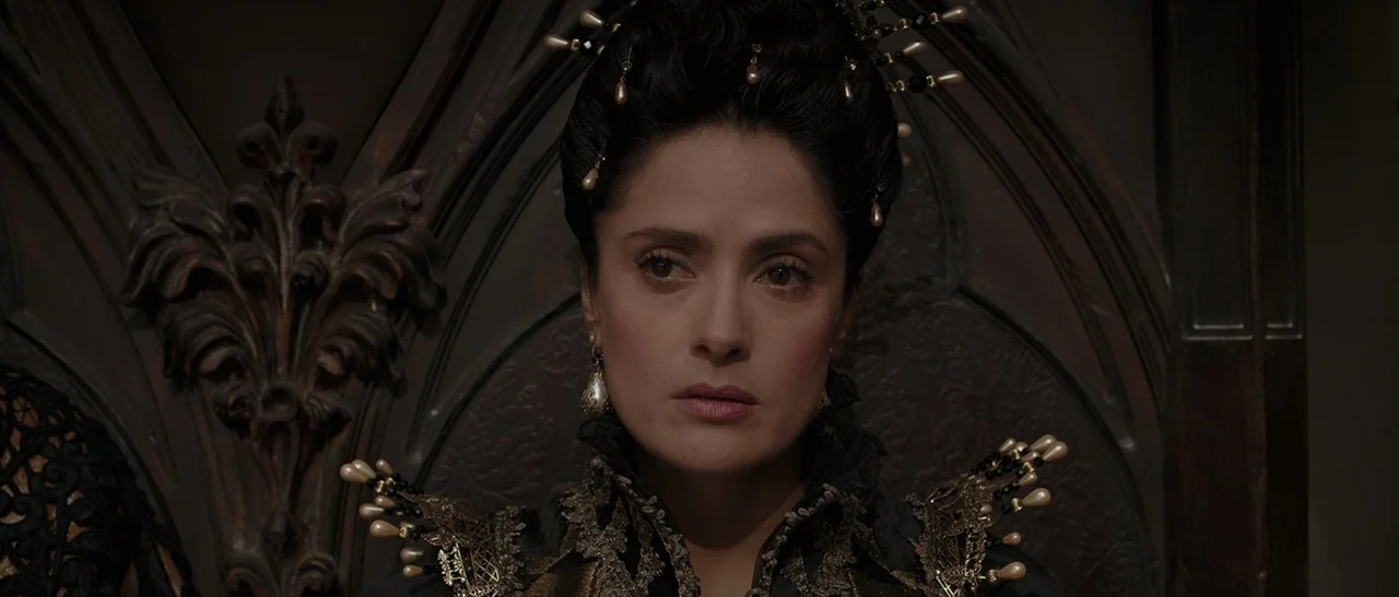 Tale of Tales (2015) movie still: closeup — A woman sits before a dark, intricately carved wooden backdrop in a dim room. She wears elaborate…