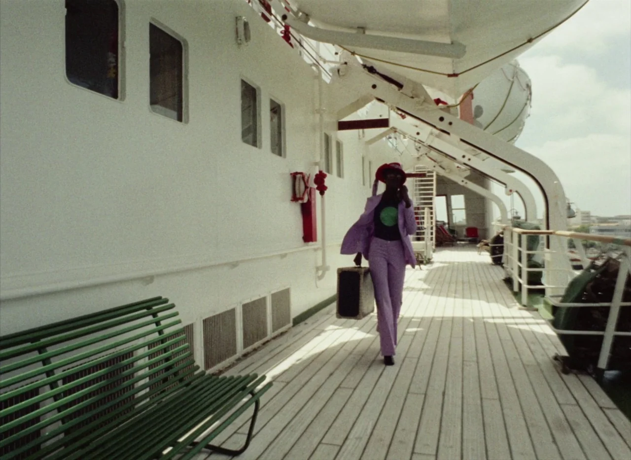 Touki Bouki (1973) movie still: fullbody — A white ship deck features a long wooden floor and green slatted benches. A woman in a purple suit walks…