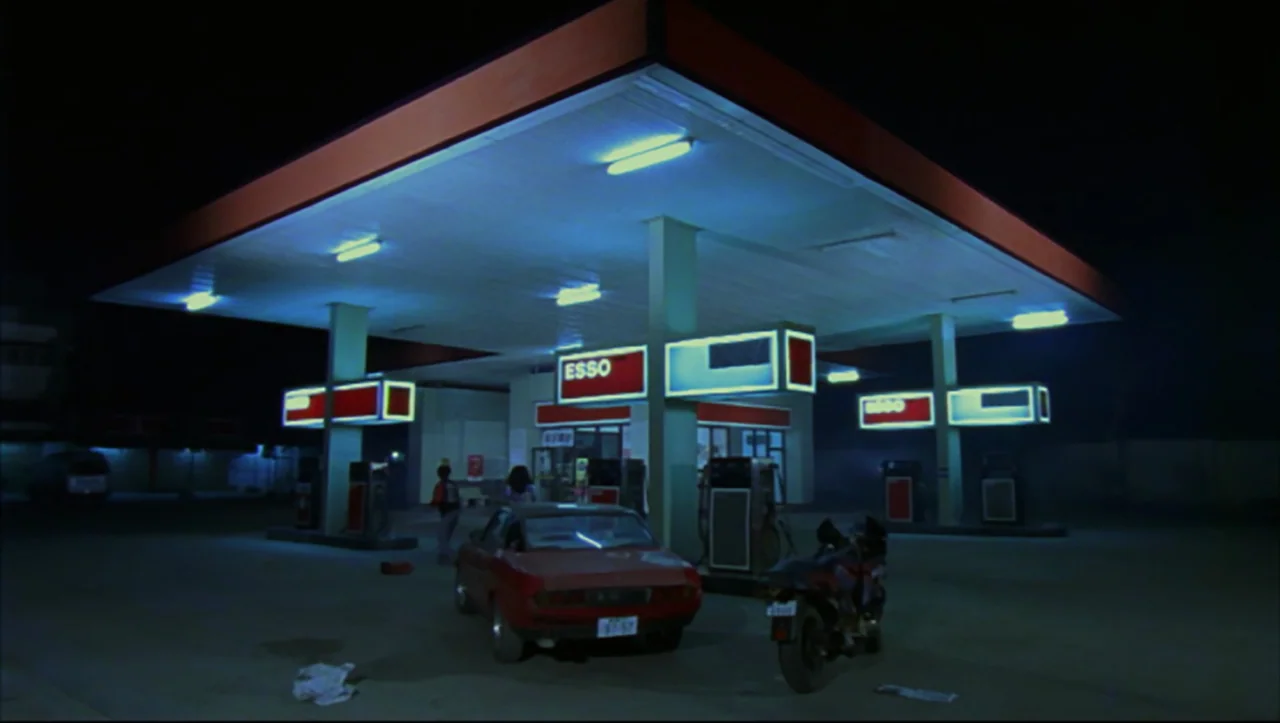 Wild Zero (1999) movie still: wide — A night view of an Esso gas station with a large illuminated overhead canopy. A red car and a motorcycle are…