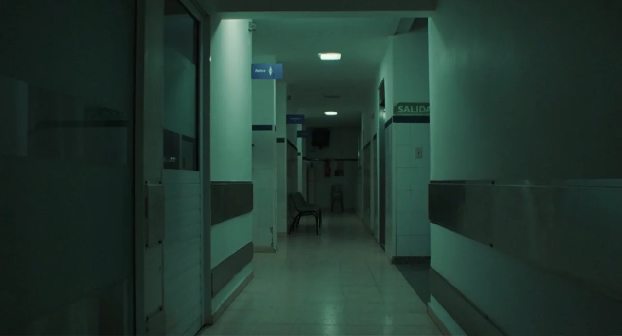 Belén (2025) movie still: establishing — A long, quiet hospital corridor with white tiled walls and overhead fluorescent lighting fixtures. The…