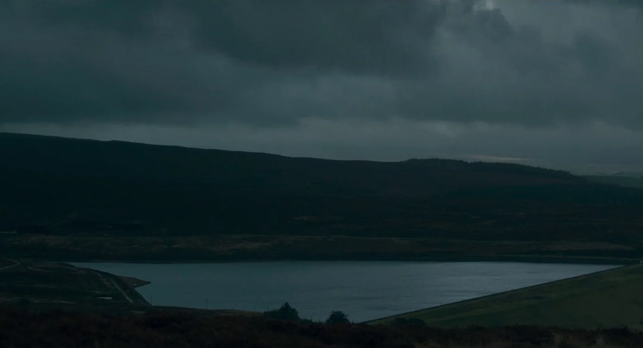 Dark River (2017) movie still: establishing — A vast, dark reservoir sits nestled between rolling, heather-covered hills under a heavy overcast sky.…