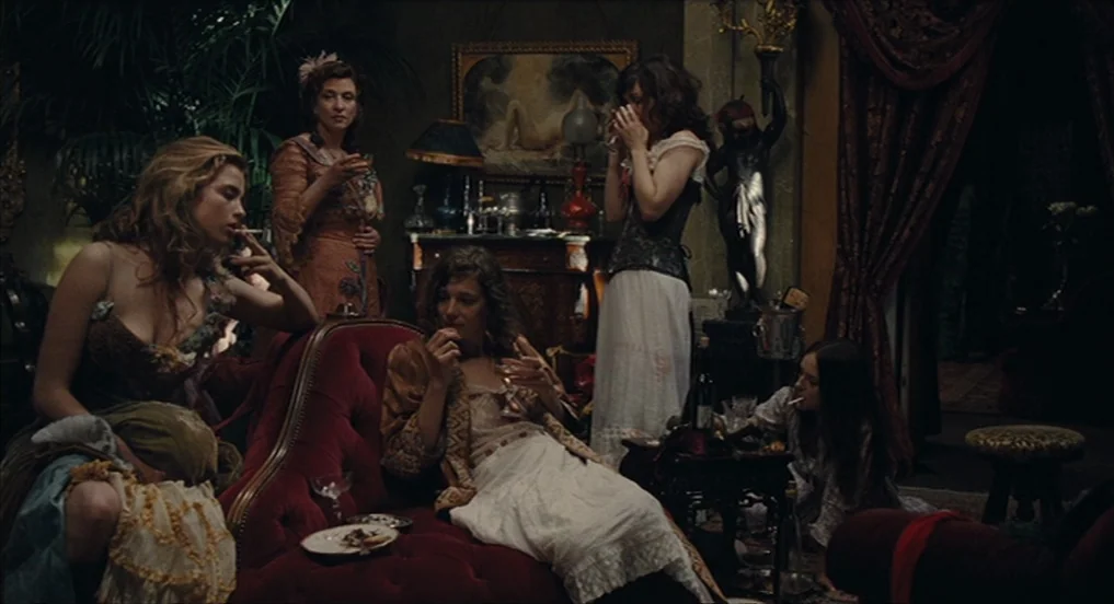 House of Tolerance (2011) movie still: medium — A dimly lit Victorian-era parlor filled with antique furniture, heavy drapes, and potted plants.…