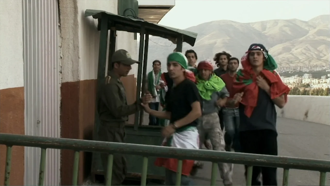 Offside (2015) movie still: medium — A paved security checkpoint with a guard hut sits before a desert mountain landscape. A group of young men with…