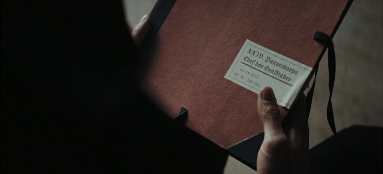 The Tank (2025) movie still: closeup — A close view of a person holding a red paper dossier with a white label. The text on the label specifies…