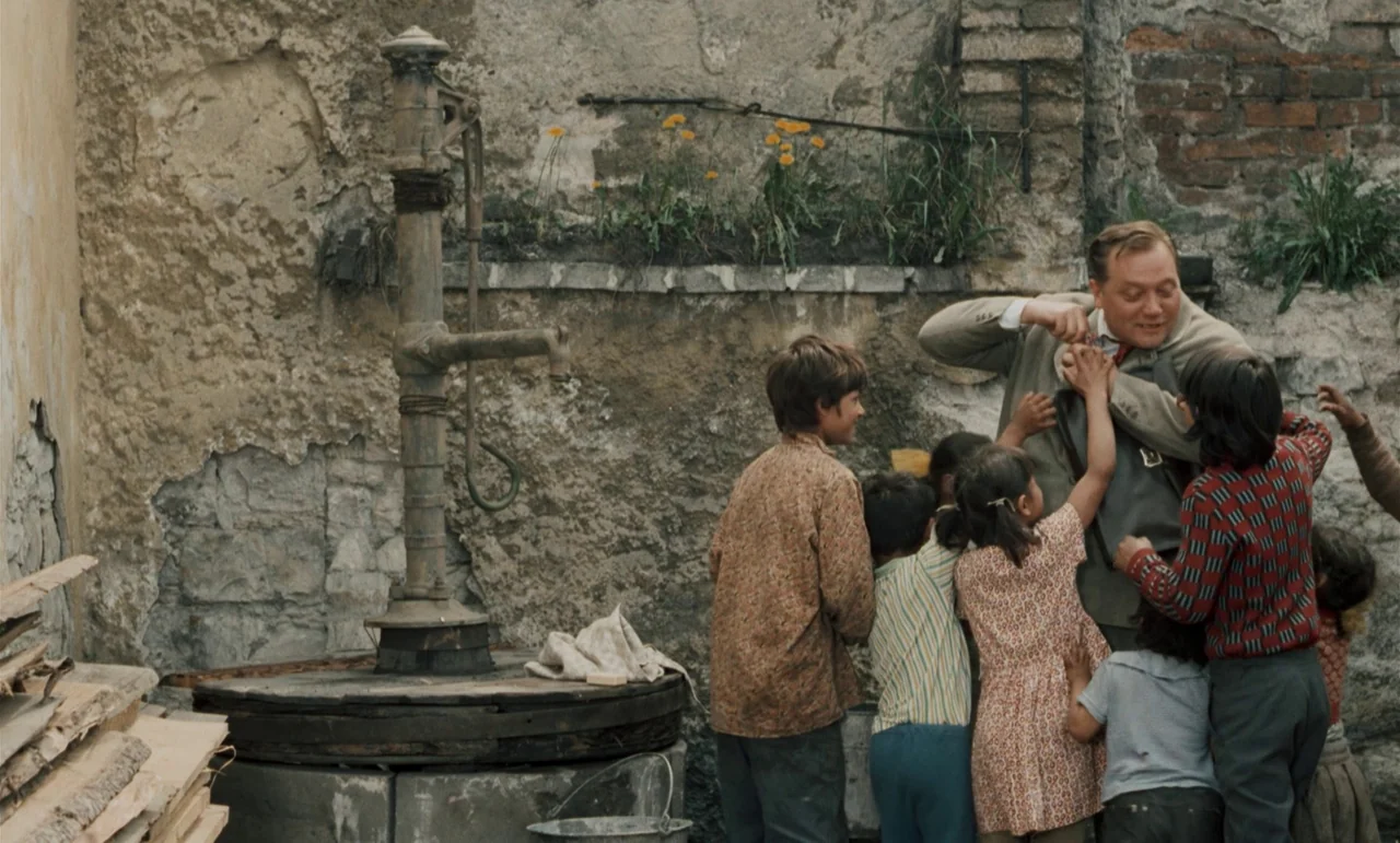Larks On A String (1969) movie still: medium — A weathered stone wall serves as a backdrop to a metal water pump. An adult man interacts with a…