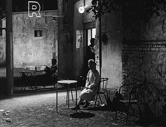 Nights of Cabiria (1957) movie still: wide — An outdoor evening scene features a woman sitting near a small shop entrance. Other people occupy…