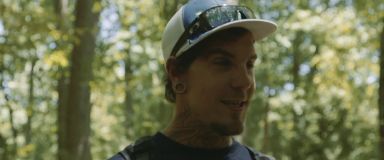 Darkness Waits (2020) movie still: closeup — A dense forest with lush green foliage and scattered patches of sunlight. A young man with tattoos and…