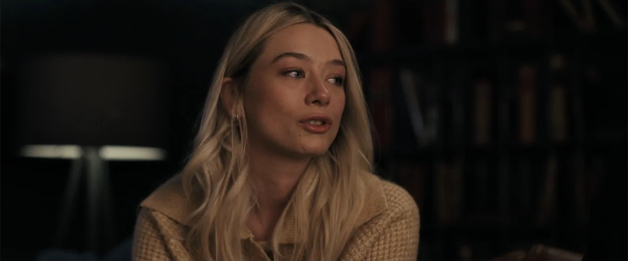 The Good Neighbor (2022) movie still: medium — A dimly lit room features bookshelves blurred in the dark background behind the subject. A blonde…