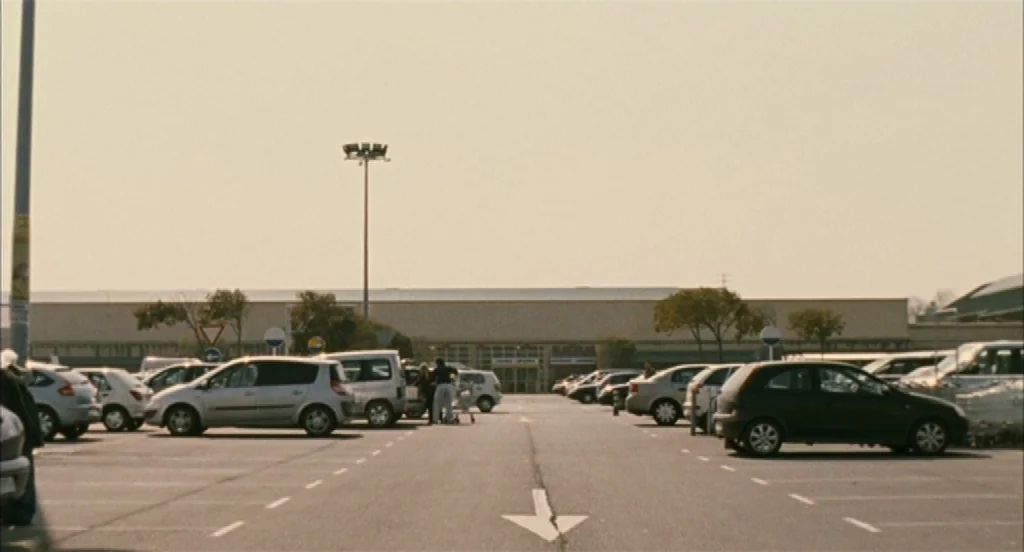 Timecrimes (2007) movie still: wide — A sprawling outdoor parking lot features rows of parked cars under a hazy sky. Two people stand near a…