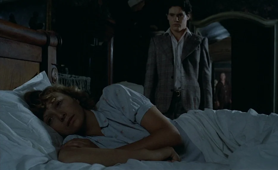 Lacombe, Lucien (1974) movie still: medium — A dimly lit room with a woman lying in a bed. A young man wearing a blazer stands near the headboard in…