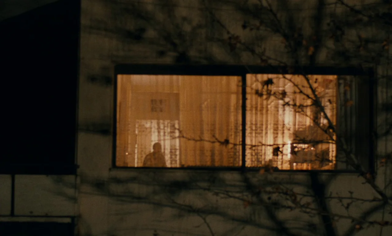 Taste of Cherry (1997) movie still: wide — The exterior of a multi-story building is dark, with tree branches casting shadows across the facade. A…