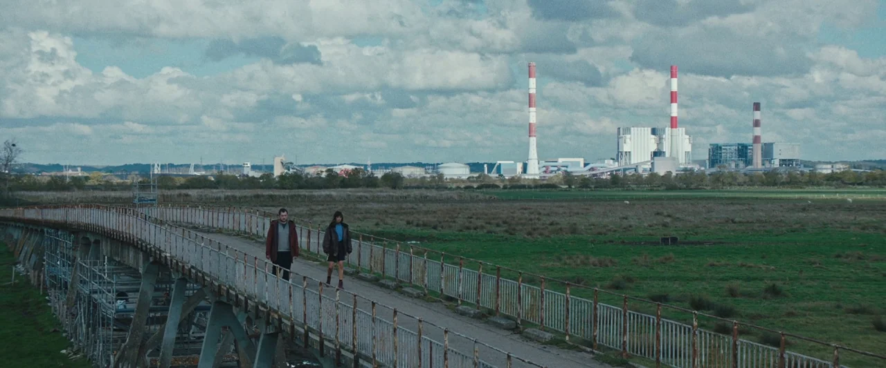 Vincent Must Die (2023) movie still: wide — A long, rusted metal walkway stretches across open green fields toward a distant power plant. A man and…