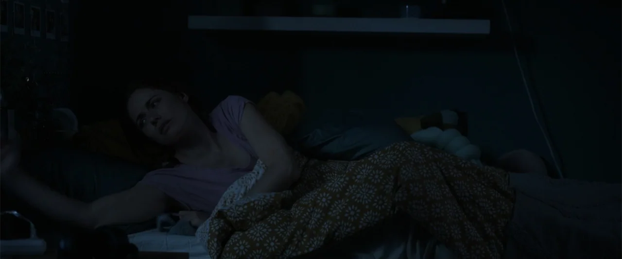 Final Destination Bloodlines (2025) movie still: medium — A dark bedroom interior features a woman lying in a bed with patterned blankets. The woman…