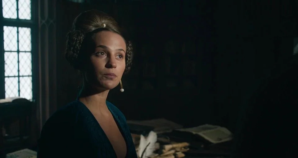 The Green Knight (2021) movie still: medium — A dark, wood-paneled room is dimly illuminated by daylight entering through a side window. A woman…