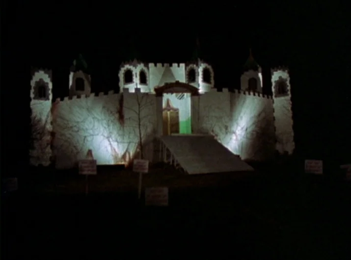 Desperate Living (1977) movie still: establishing — A miniature wooden toy castle sits in total darkness at night. The structure features turrets, a…
