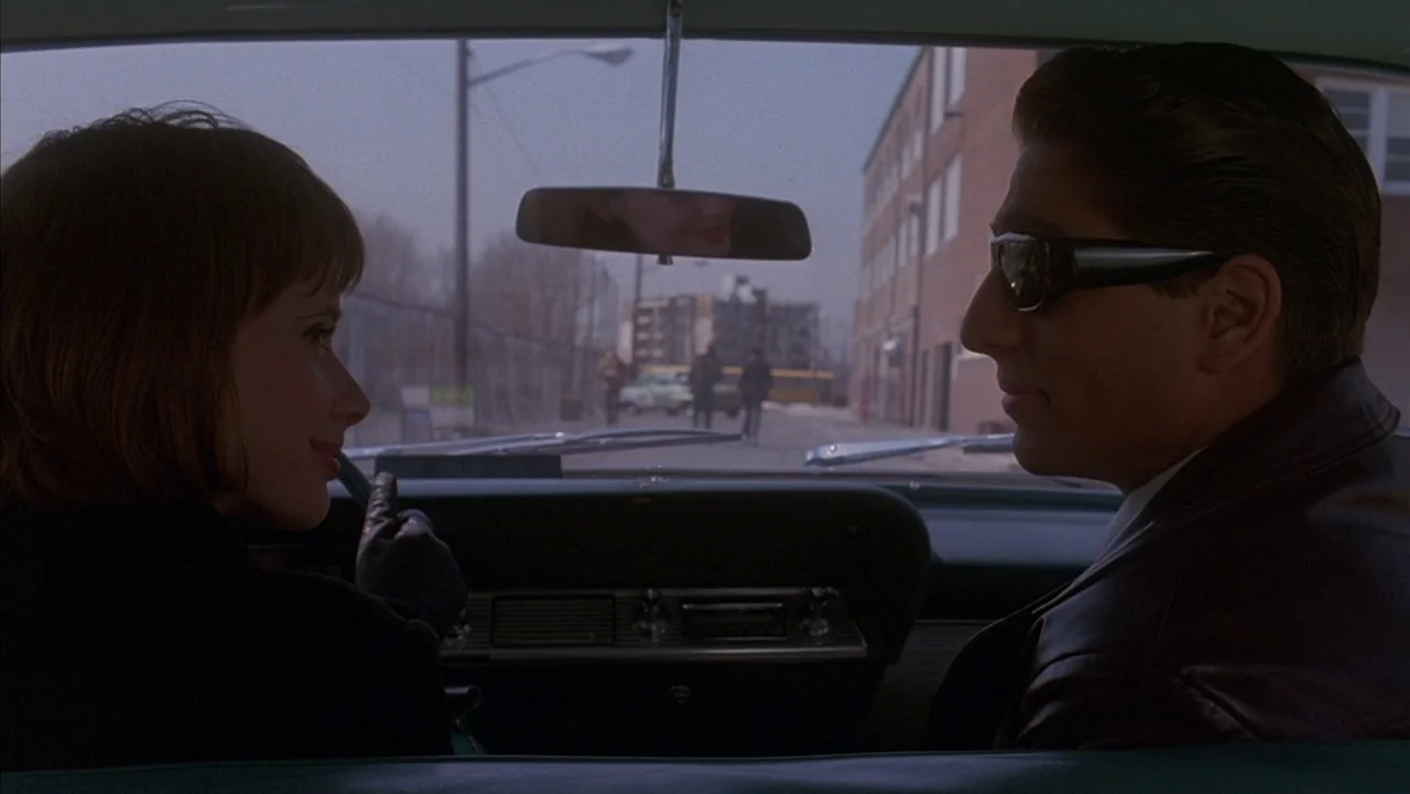 Baby It’s You (1983) movie still: medium — The interior of a mid-90s sedan parked on an urban street with brick buildings. A man in sunglasses and a…