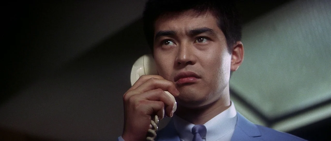 Tokyo Drifter (1966) movie still: closeup — An interior space at night with dark surroundings. A young man speaks into a white rotary telephone…