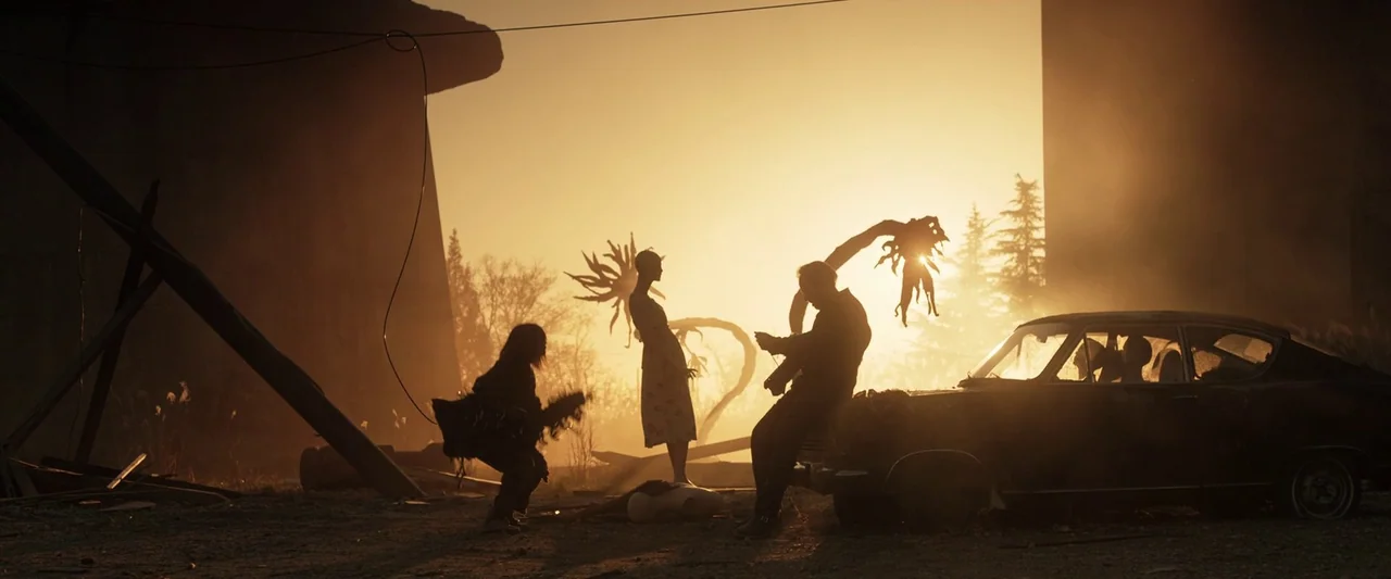 Prisoners of the Ghostland (2021) movie still: wide — A desolate, hazy open space containing debris and a wrecked car at sunset. Three figures…