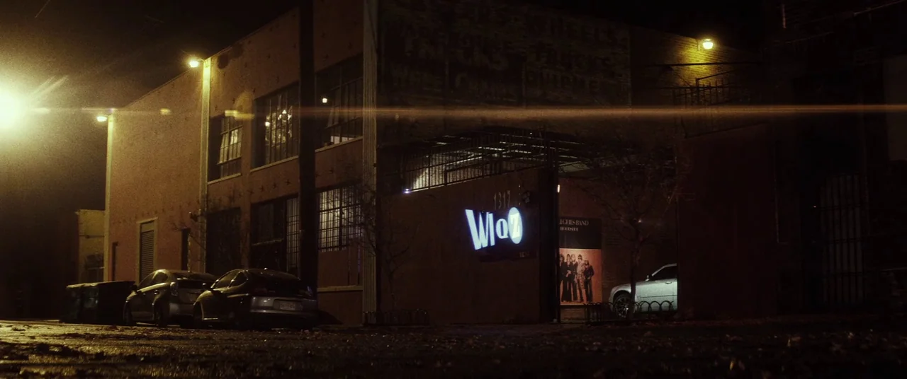 Lords Of Salem (2013) movie still: wide — A dark brick building at night features a glowing neon sign above a gated entrance. Two parked cars sit on…
