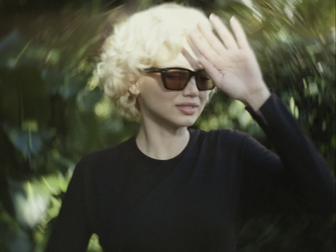 Blonde (2022) movie still: medium — A person stands outdoors against a background of out-of-focus green foliage. The blonde subject holds a hand up…
