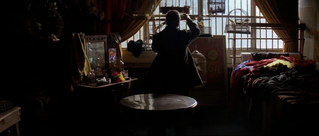 The Prestige (2006) movie still: wide — A cluttered room with numerous birdcages and heavy fabric curtains hung by a window. The man stands facing…