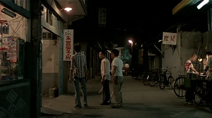 A Time To Love and A Time To Die (1985) movie still: wide — A quiet urban night scene in a narrow alleyway with various shops. Three men stand…