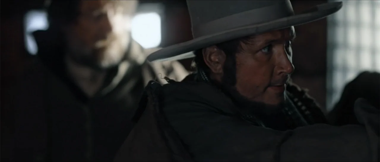 Out of Liberty (2019) movie still: medium — A dimly lit room with rough wooden walls and small window openings. A man wearing a wide-brimmed hat…