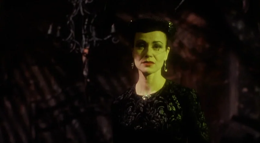 Dust Devil (1992) movie still: medium — A dark, dimly lit castle interior with indistinct textures in the shadows. A woman with green-tinted skin…