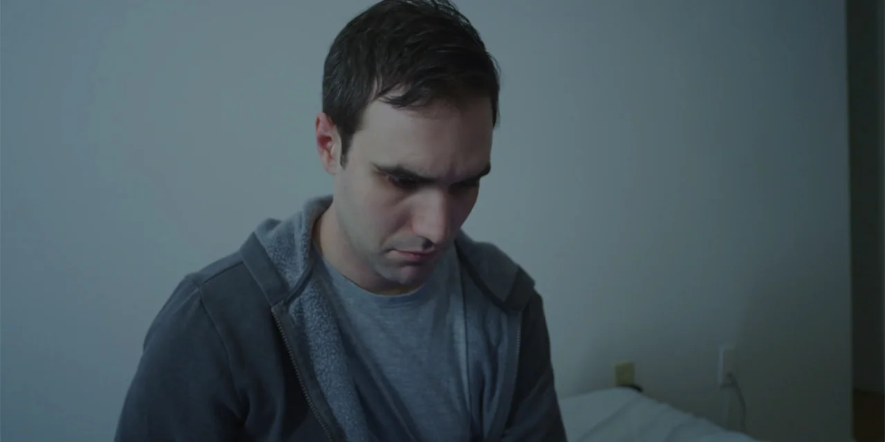 Lobos (2019) movie still: medium — A plain interior room with neutral walls and a bed edge visible. A man in a grey hoodie sits alone while looking…