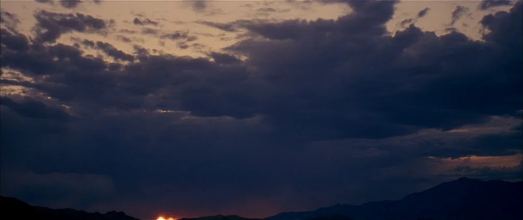 Gerry (2002) movie still: establishing — A vast mountain desert landscape is covered by a dense layer of dark clouds. The final sliver of sunset…