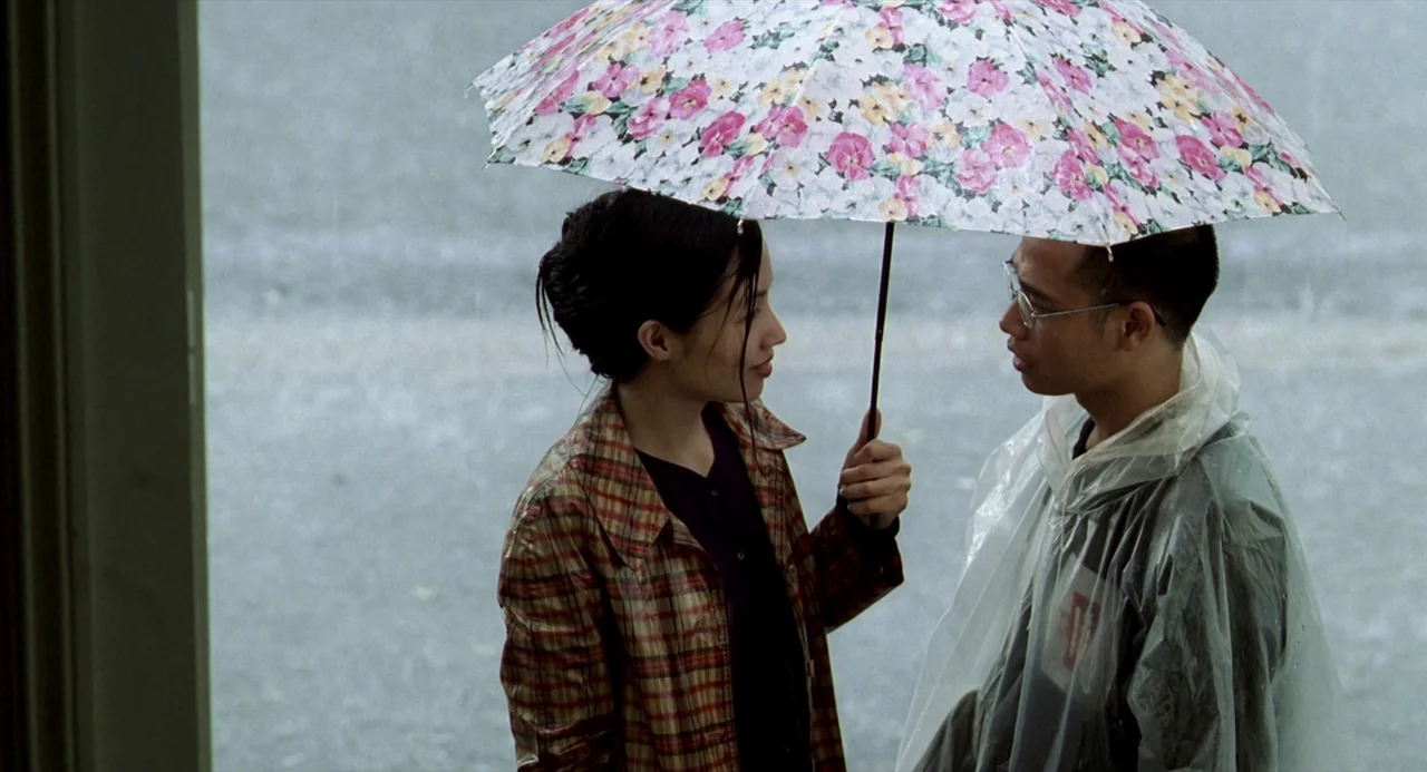 The Vertical Ray of the Sun (2000) movie still: medium — An outdoor sidewalk during a heavy rainstorm with visible precipitation falling. A young…