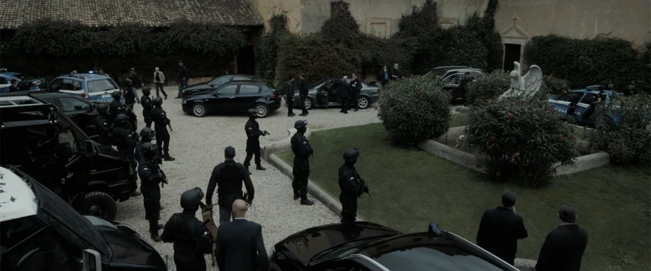 The Equalizer 3 (2023) movie still: wide — A stone-walled courtyard features gravel paths, a central grass patch, and an angel statue. Numerous…
