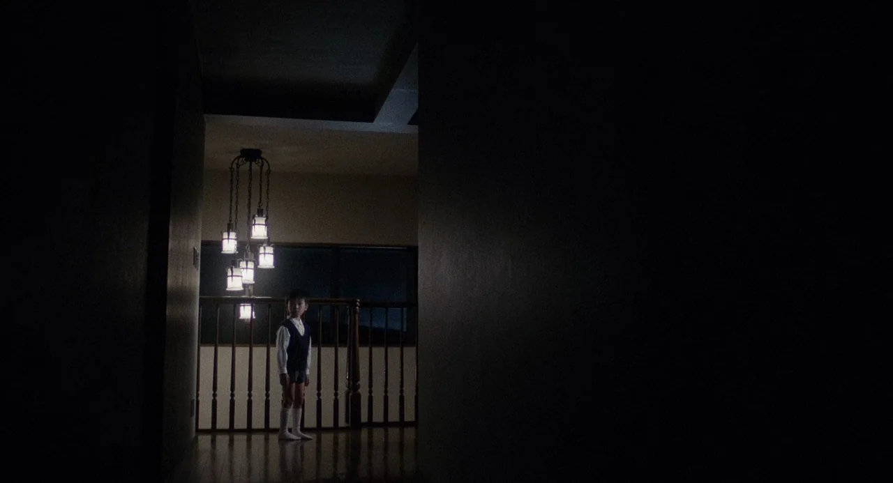 Ringu (1998) movie still: wide — A dark, empty domestic corridor opens into a brightly lit area with a wooden railing. A young child stands solitary…
