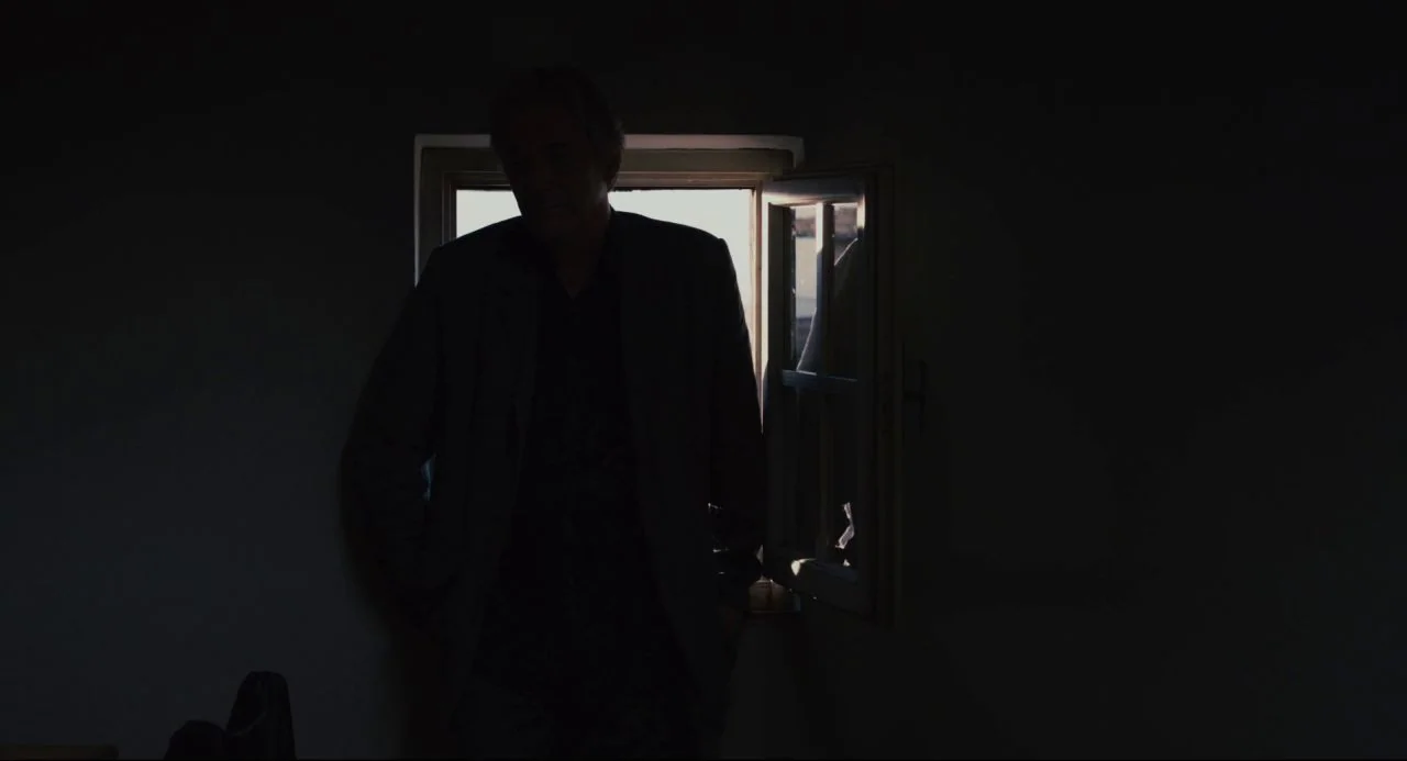 Certified Copy (2010) movie still: medium — A dark, dimly lit room features a subject standing before an open window. The man is positioned against…