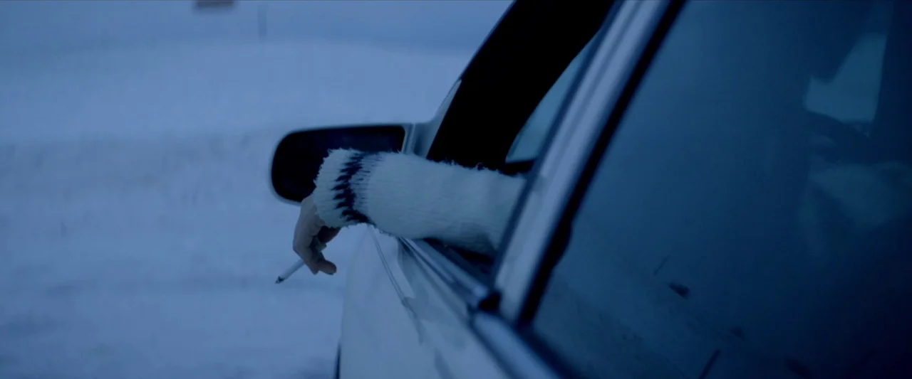 The Unknown Country (2022) movie still: closeup — A white car side mirror and window frame against a flat, snow-covered landscape. A person's hand…