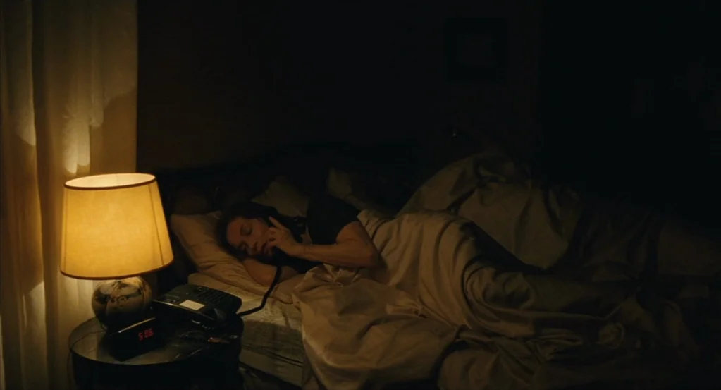 Things to Come (2016) movie still: medium — A dark bedroom features a wooden side table with a lit lamp and telephone. A woman lies under covers in…