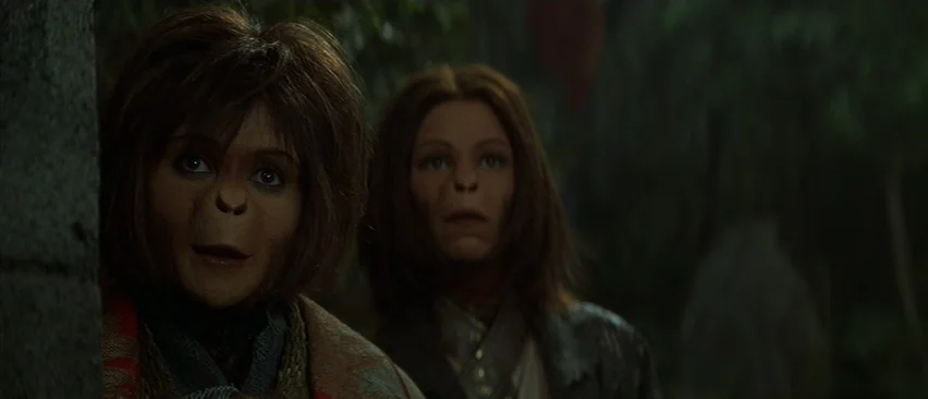 Planet of the Apes (2001) movie still: medium — Two simian characters stand amidst dense green foliage in a natural forest environment. They look…