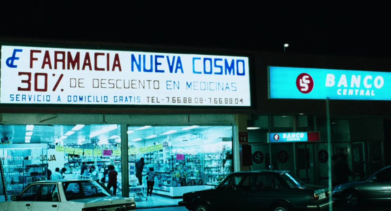 Amores Perros (2000) movie still: wide — A nighttime view of a commercial strip featuring a brightly lit pharmacy and bank. The storefronts have…