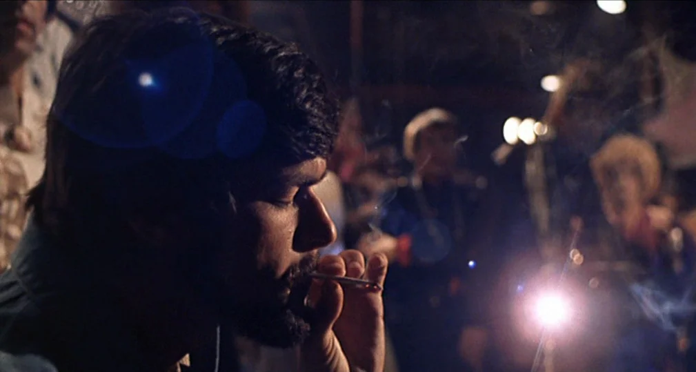 Midnight Cowboy (1969) movie still: closeup — A dimly lit, crowded space with blurred silhouettes of people standing in the back. A young man in…
