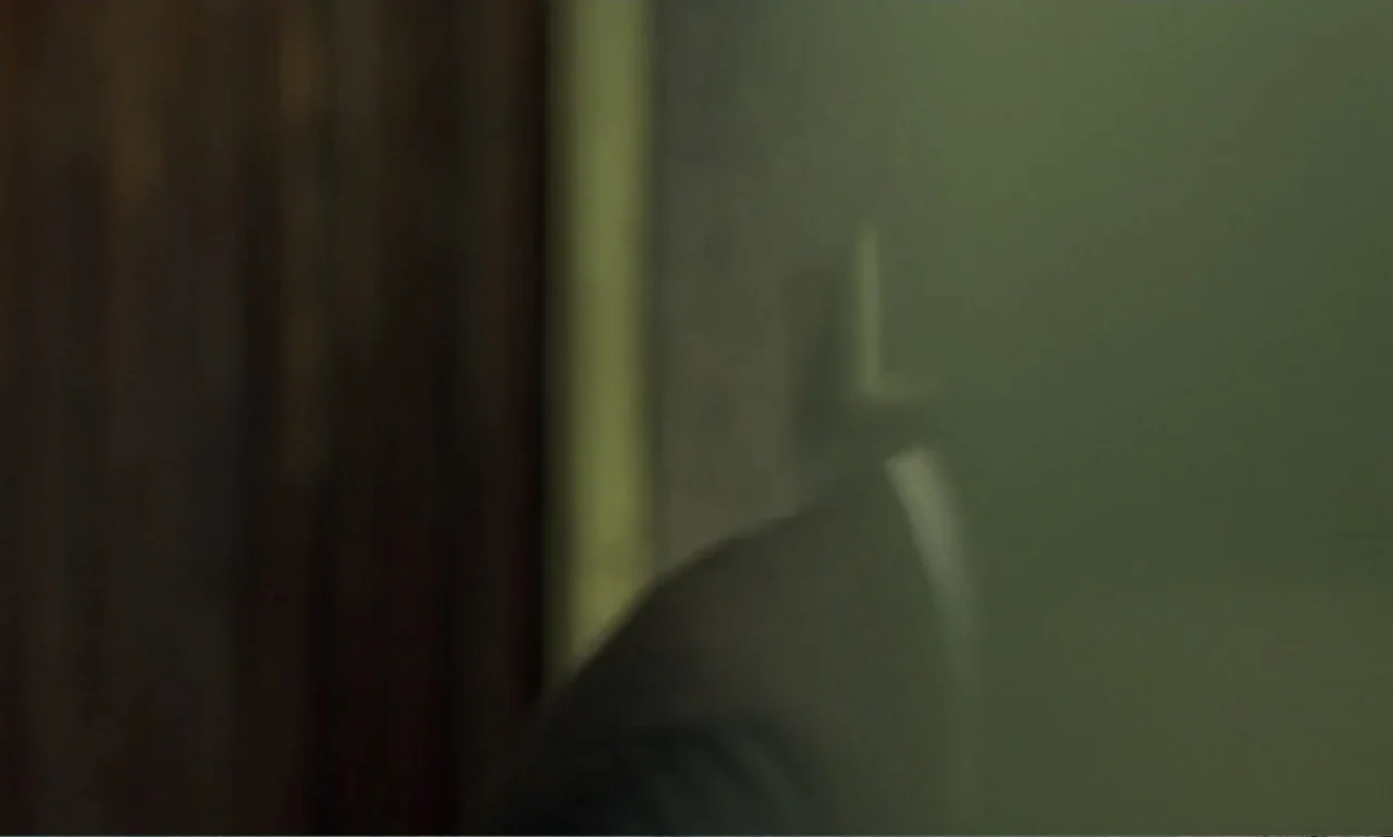 In the Mood for Love (2000) movie still: closeup — A dimly lit indoor space features a dark wall and a green-lit area. A person wearing a dark suit…
