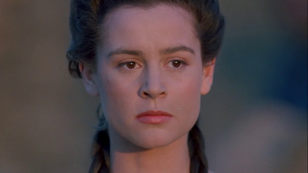 Army of Darkness (1992) movie still: closeup — A woman stands in a blurred outdoor field during the golden hour. She faces forward with a neutral…