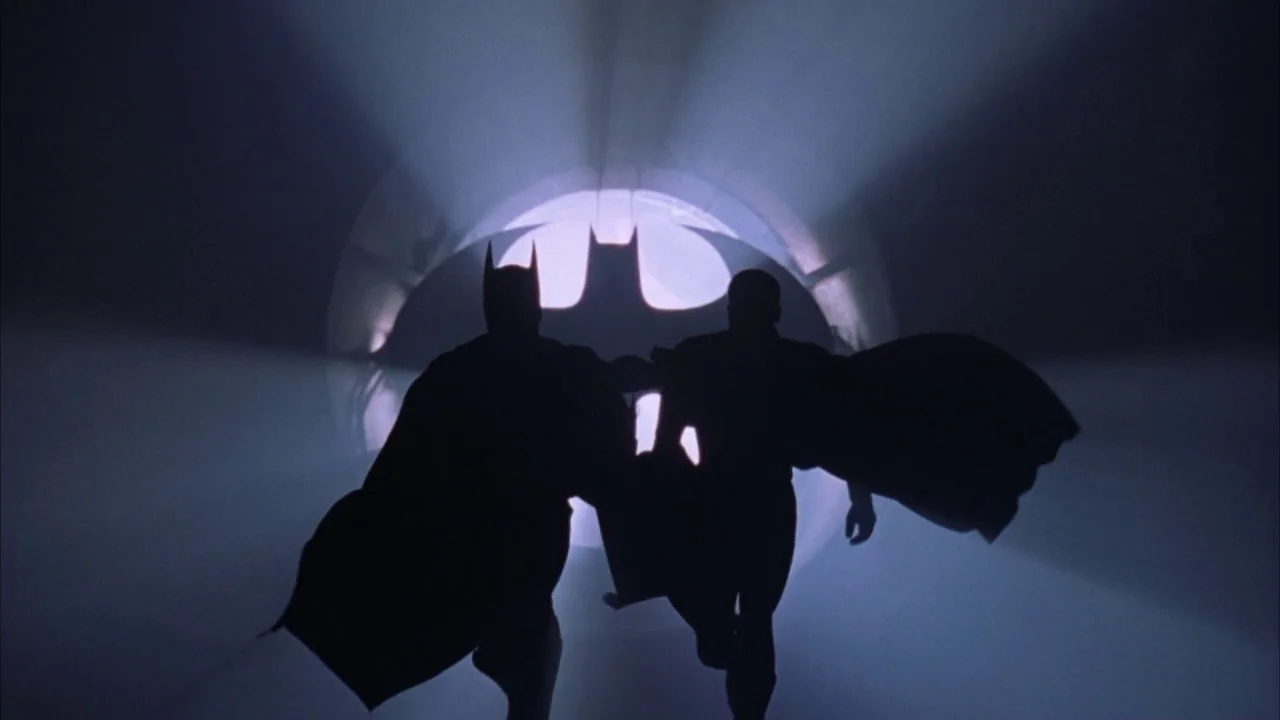 Batman Forever (1995) movie still: fullbody — A long, dark concrete tunnel is illuminated by a bright, circular projected bat symbol. Two caped…