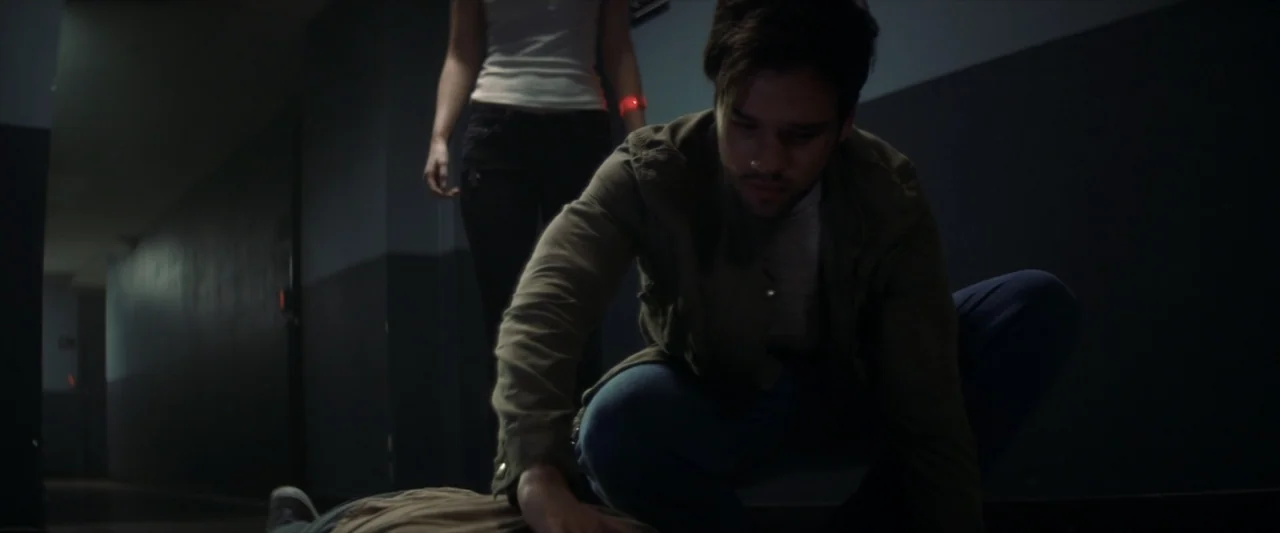 Tell Me How I Die (2016) movie still: medium — A dark, modern interior corridor with grey walls and indistinct background details. A man kneels on…